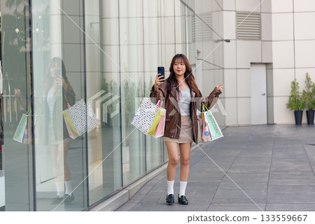 Young Asian woman enjoys upbeat shopping lifestyle, holding colorful fashion shopping bags, posing with smartphone near modern mall building, city street setting. Young Asian woman enjoys upbeat shopping lifestyle, holding colorful fashion shopping bags, posing with smartphone near modern mall building, city street setting. 133559667