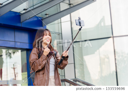 Smiling Asian woman, people enjoy shopping lifestyle fashion, posing with phone selfie stick for social media outside modern city mall building, capturing travel memory. 133559696