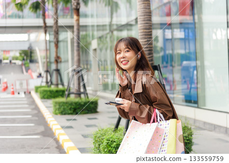 Asian woman enjoys shopping lifestyle, holding colorful fashion bags near modern city mall, smiling, looking away, street with palm trees, people enjoying urban retail during black friday season. Asian woman enjoys shopping lifestyle, holding colorful fashion bags near modern city mall, smiling, looking away, street with palm trees, people enjoying urban retail during black friday season. 133559759