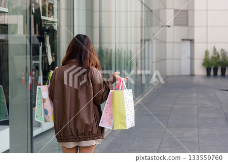 Asian woman enjoys shopping carries many colorful fashion bags walking past modern city mall building exuding modern lifestyle for people during black friday shop sales event. Asian woman enjoys shopping carries many colorful fashion bags walking past modern city mall building exuding modern lifestyle for people during black friday shop sales event. 133559760