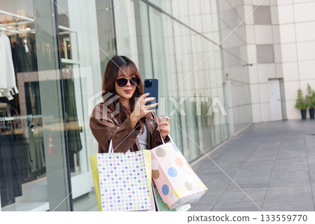 Shopping woman enjoys bags lifestyle, fashion buyer near mall outside, people gathering for black friday sales, Asian woman posing with phone and sunglasses. 133559770