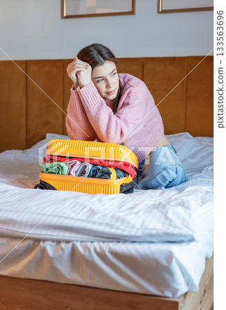 Woman packing suitcase for vacation trip while sitting on bed Woman packing suitcase for vacation trip while sitting on bed 133563696