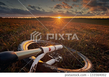 A bicycle wheel against the background of a meadow and a bright sunset sky with the sun. Travel and adventure 133564002