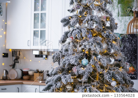 Christmas decor in white kitchen, mess dishes, contemporary interior with a snowy Christmas tree and gold fairy lights. New Year, Christmas mood, cozy home. 133564028