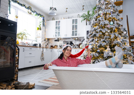 Happy woman in Santa hat is lying resting in New bathtub under Christmas tree as New Year's gift in interior of decorated kitchen. Interior renovation and design, construction and home improvement Happy woman in Santa hat is lying resting in New bathtub under Christmas tree as New Year's gift in interior of decorated kitchen. Interior renovation and design, construction and home improvement 133564031
