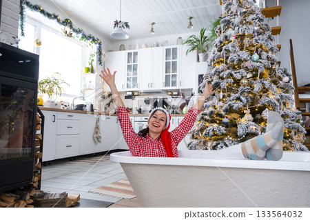 Happy woman in Santa hat is lying resting in New bathtub under Christmas tree as New Year's gift in interior of decorated kitchen. Interior renovation and design, construction and home improvement Happy woman in Santa hat is lying resting in New bathtub under Christmas tree as New Year's gift in interior of decorated kitchen. Interior renovation and design, construction and home improvement 133564032