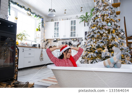 Happy woman in Santa hat is lying resting in New bathtub under Christmas tree as New Year's gift in interior of decorated kitchen. Interior renovation and design, construction and home improvement Happy woman in Santa hat is lying resting in New bathtub under Christmas tree as New Year's gift in interior of decorated kitchen. Interior renovation and design, construction and home improvement 133564034