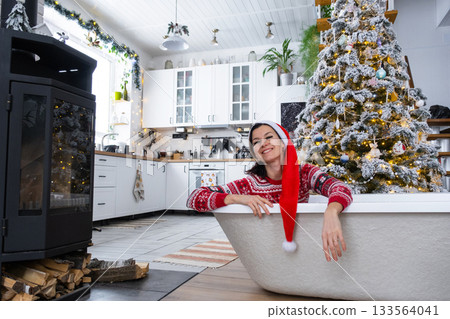 Happy woman in Santa hat is lying resting in New bathtub under Christmas tree as New Year's gift in interior of decorated kitchen. Interior renovation and design, construction and home improvement Happy woman in Santa hat is lying resting in New bathtub under Christmas tree as New Year's gift in interior of decorated kitchen. Interior renovation and design, construction and home improvement 133564041