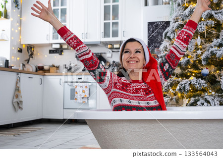 Happy woman in Santa hat is lying resting in New bathtub under Christmas tree as New Year's gift in interior of decorated kitchen. Interior renovation and design, construction and home improvement Happy woman in Santa hat is lying resting in New bathtub under Christmas tree as New Year's gift in interior of decorated kitchen. Interior renovation and design, construction and home improvement 133564043