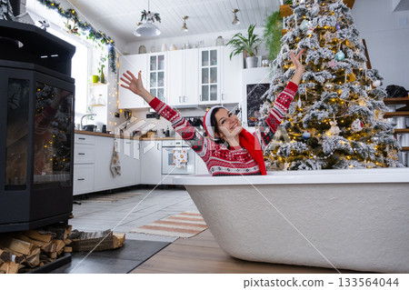 Happy woman in Santa hat is lying resting in New bathtub under Christmas tree as New Year's gift in interior of decorated kitchen. Interior renovation and design, construction and home improvement 133564044