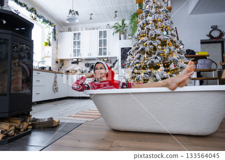 Happy woman in Santa hat is lying resting in New bathtub under Christmas tree as New Year's gift in interior of decorated kitchen. Interior renovation and design, construction and home improvement 133564045