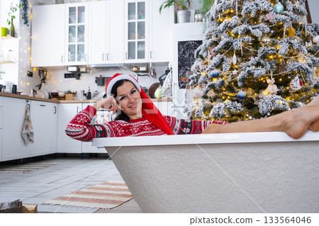 Happy woman in Santa hat is lying resting in New bathtub under Christmas tree as New Year's gift in interior of decorated kitchen. Interior renovation and design, construction and home improvement 133564046