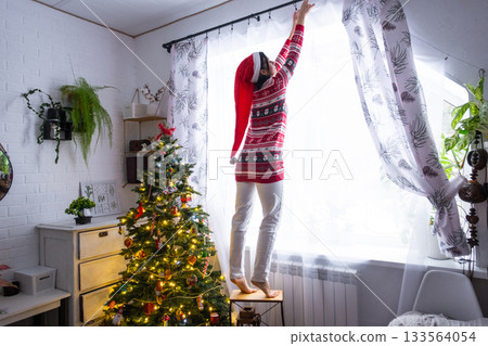 A woman in a Santa hat hangs curtains in the bedroom on the skylights with a Christmas tree and Christmas decorations, preparing for the New Year 133564054