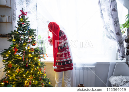 A woman in a Santa hat hangs curtains in the bedroom on the skylights with a Christmas tree and Christmas decorations, preparing for the New Year A woman in a Santa hat hangs curtains in the bedroom on the skylights with a Christmas tree and Christmas decorations, preparing for the New Year 133564059