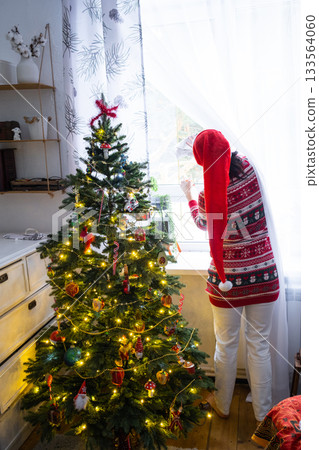 A woman in a Santa hat hangs curtains in the bedroom on the skylights with a Christmas tree and Christmas decorations, preparing for the New Year 133564060