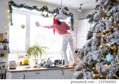 A woman in a Santa hat cleans a kitchen window with Christmas decorations, preparing for the New Year, decorating and tidying up the house 133564065