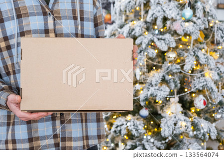 A courier with a parcel box in the kitchen with white Christmas decorations and a Christmas tree. Gift delivery for New Year and Christmas, courier service 133564074