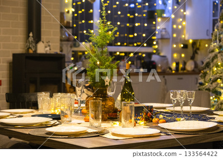 Festive interior of house is decorated for Christmas and New Year in loft style with black stove, fireplace, Christmas tree. Warm studio room with set table, burning wood, cozy and heating of home 133564422