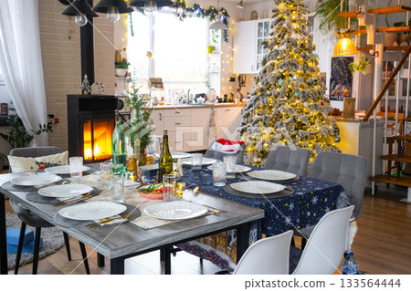 Festive interior of house is decorated for Christmas and New Year in loft style with black stove, fireplace, Christmas tree. Warm studio room with set table, burning wood, cozy and heating of home Festive interior of house is decorated for Christmas and New Year in loft style with black stove, fireplace, Christmas tree. Warm studio room with set table, burning wood, cozy and heating of home 133564444