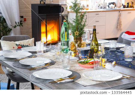 A festive table set for guests for Christmas and New Year, a Christmas tree in the living room, plates, bottles, glasses, potted plants, fairy lights 133564445