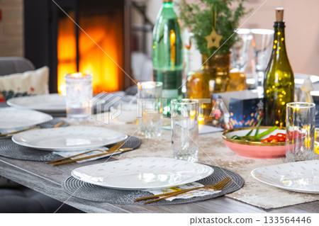 A festive table set for guests for Christmas and New Year, a Christmas tree in the living room, plates, bottles, glasses, potted plants, fairy lights A festive table set for guests for Christmas and New Year, a Christmas tree in the living room, plates, bottles, glasses, potted plants, fairy lights 133564446