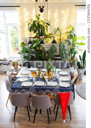 A festive table set for guests for Christmas and New Year, a Christmas tree in the living room, plates, bottles, glasses, potted plants, fairy lights A festive table set for guests for Christmas and New Year, a Christmas tree in the living room, plates, bottles, glasses, potted plants, fairy lights 133564456
