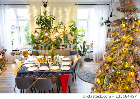 A festive table set for guests for Christmas and New Year, a Christmas tree in the living room, plates, bottles, glasses, potted plants, fairy lights 133564457