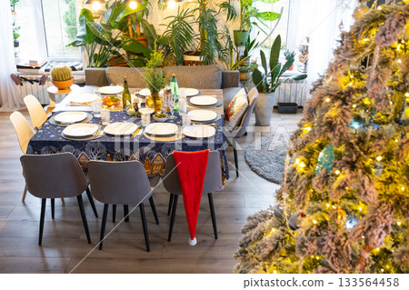 A festive table set for guests for Christmas and New Year, a Christmas tree in the living room, plates, bottles, glasses, potted plants, fairy lights 133564458