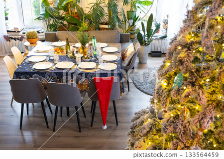 A festive table set for guests for Christmas and New Year, a Christmas tree in the living room, plates, bottles, glasses, potted plants, fairy lights 133564459