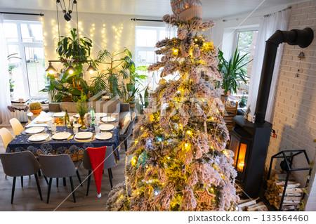 Festive interior of house is decorated for Christmas and New Year in loft style with black stove, fireplace, Christmas tree. Warm studio room with set table, burning wood, cozy and heating of home 133564460