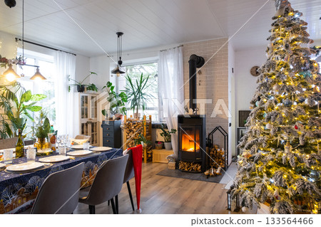 Festive interior of house is decorated for Christmas and New Year in loft style with black stove, fireplace, Christmas tree. Warm studio room with set table, burning wood, cozy and heating of home 133564466