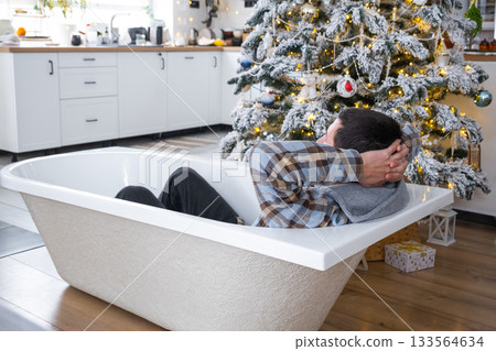 Man is lying resting in New bathtub under Christmas tree as New Year's gift in interior of white decorated kitchen. Interior renovation and design, construction and home improvement 133564634