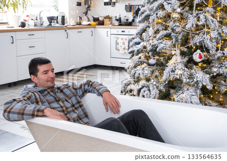 Man is lying resting in New bathtub under Christmas tree as New Year's gift in interior of white decorated kitchen. Interior renovation and design, construction and home improvement 133564635