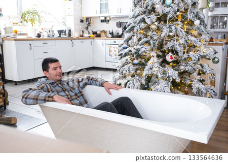 Man is lying resting in New bathtub under Christmas tree as New Year's gift in interior of white decorated kitchen. Interior renovation and design, construction and home improvement 133564636