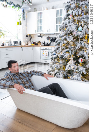 Man is lying resting in New bathtub under Christmas tree as New Year's gift in interior of white decorated kitchen. Interior renovation and design, construction and home improvement 133564637