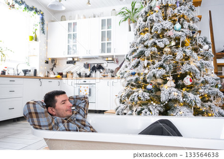 Man is lying resting in New bathtub under Christmas tree as New Year's gift in interior of white decorated kitchen. Interior renovation and design, construction and home improvement 133564638