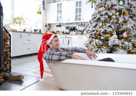 Man in Santa hat is lying resting in New bathtub under Christmas tree as New Year's gift in interior of white decorated kitchen. Interior renovation and design, construction and home improvement 133564641