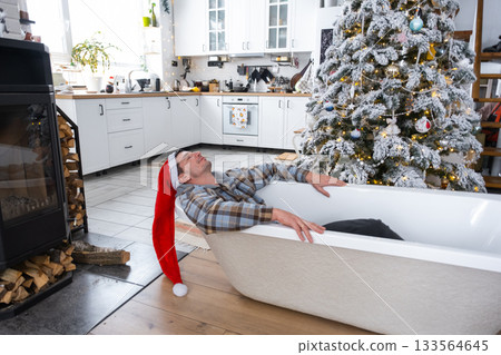 Man in Santa hat is lying resting in New bathtub under Christmas tree as New Year's gift in interior of white decorated kitchen. Interior renovation and design, construction and home improvement 133564645