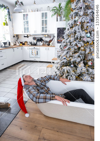 Man in Santa hat is lying resting in New bathtub under Christmas tree as New Year's gift in interior of white decorated kitchen. Interior renovation and design, construction and home improvement Man in Santa hat is lying resting in New bathtub under Christmas tree as New Year's gift in interior of white decorated kitchen. Interior renovation and design, construction and home improvement 133564646