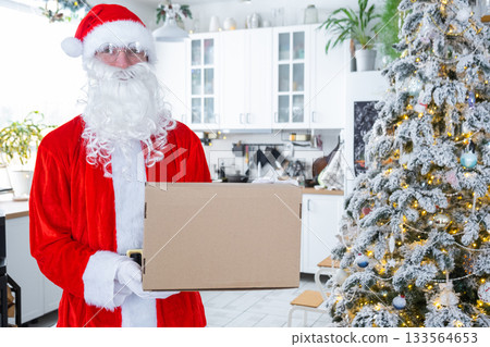 Courier Dressed as Santa with parcel box in the kitchen with white Christmas decorations and a Christmas tree. Gift delivery for New Year and Christmas, courier service 133564653