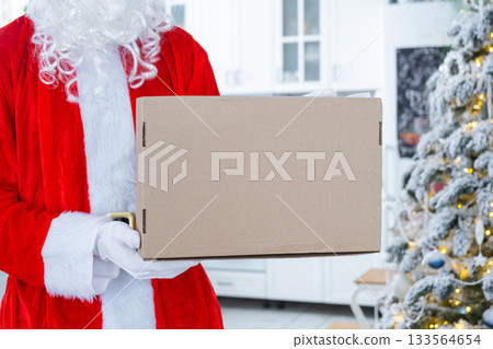 Courier Dressed as Santa with parcel box in the kitchen with white Christmas decorations and a Christmas tree. Gift delivery for New Year and Christmas, courier service Courier Dressed as Santa with parcel box in the kitchen with white Christmas decorations and a Christmas tree. Gift delivery for New Year and Christmas, courier service 133564654