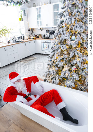 Santa is lying resting in New bathtub under festive Christmas tree as New Year's gift in interior of white modern decorated kitchen. Interior renovation and design, construction and home improvement Santa is lying resting in New bathtub under festive Christmas tree as New Year's gift in interior of white modern decorated kitchen. Interior renovation and design, construction and home improvement 133564664