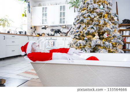 Santa is lying resting in New bathtub under festive Christmas tree as New Year's gift in interior of white modern decorated kitchen. Interior renovation and design, construction and home improvement Santa is lying resting in New bathtub under festive Christmas tree as New Year's gift in interior of white modern decorated kitchen. Interior renovation and design, construction and home improvement 133564666