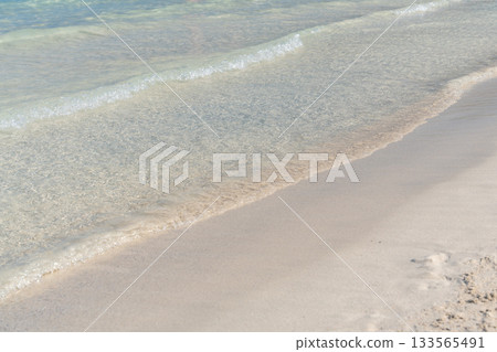 A small wave gently reaches the sandy beach, showcasing the clarity of pale turquoise sea water. The simple natural composition reflects calmness and a serene coastal moment. A small wave gently reaches the sandy beach, showcasing the clarity of pale turquoise sea water. The simple natural composition reflects calmness and a serene coastal moment. 133565491