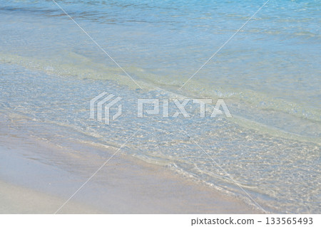 Gentle waves wash over a shallow sandy shoreline, revealing the clarity of turquoise sea water. The soft texture and natural light convey a peaceful beach atmosphere 133565493