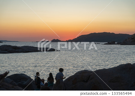 Silhouettes of people sitting on coastal rocks while watching a vibrant sunset over the calm sea. Warm orange light reflects on the water, capturing a peaceful evening moment in a natural seaside Silhouettes of people sitting on coastal rocks while watching a vibrant sunset over the calm sea. Warm orange light reflects on the water, capturing a peaceful evening moment in a natural seaside 133565494