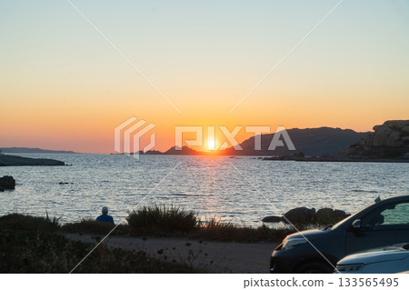 A vivid sunset descends over the sea, illuminating the coastline with warm orange tones. A parked car on the shore adds a touch of everyday life to the tranquil seaside landscape 133565495