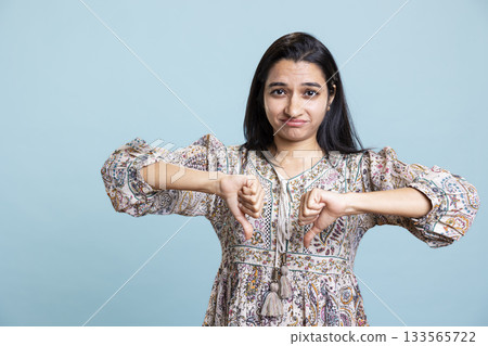 Indian frustrated woman shows thumbs down gesture in the studio, expressing disapproval and dissatisfaction. Negative skeptical person gives her bad opinion, being in disagreement. 133565722