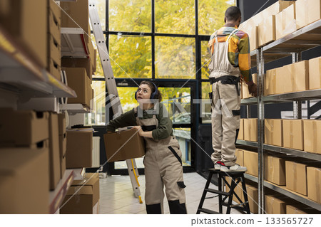 Small depot employee moves cardboard boxes with music on headset, wearing coveralls and preparing parcels for shipment. Arranging goods on shelving units for easy e-shop distribution. Small depot employee moves cardboard boxes with music on headset, wearing coveralls and preparing parcels for shipment. Arranging goods on shelving units for easy e-shop distribution. 133565727