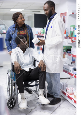 Pharmacist counseling a customer with disability on treatment options, discussing about medication dosage to support patient health. Professional medical staff showing drugstore pills. 133565728